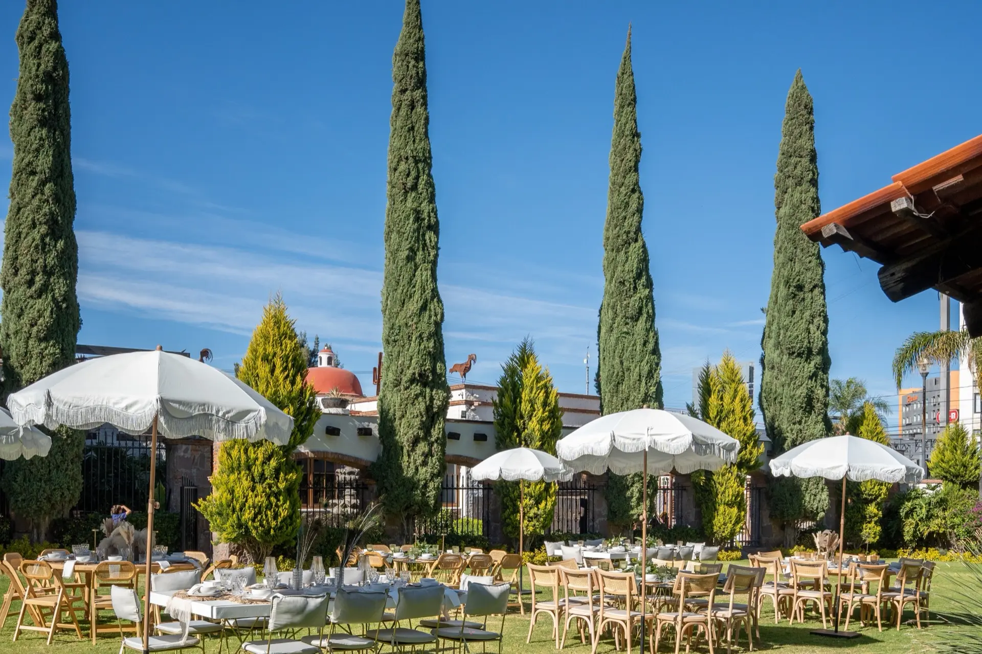 Jardín de eventos con mesas y sombrillas blancas entre cipreses