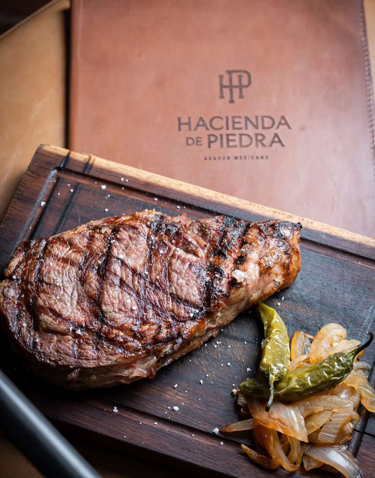 Rib eye a la parrilla con sal en escamas, chile toreado y cebollas, sobre tabla de madera junto a la carta de Hacienda de Piedra